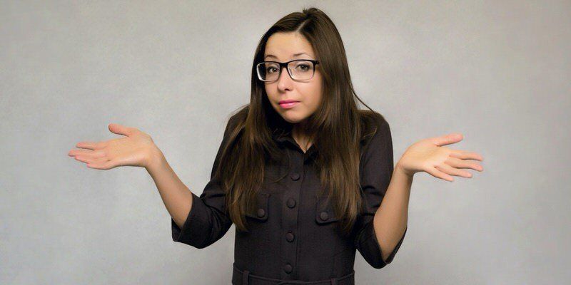 Woman with long dark hair and glasses shrugging with both hands raised against a plain gray background