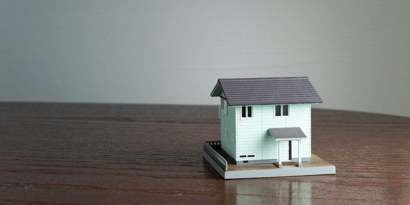 Miniature white house model with a gray roof on a wooden surface against a plain background