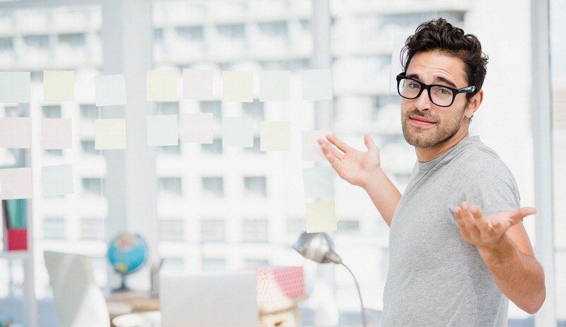 Man in glasses shrugging in a bright office, with a desk and city buildings in the background.