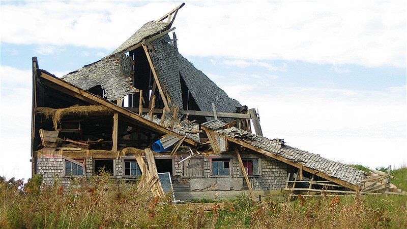 Severely damaged house with collapsed roof and broken walls in a grassy field under a cloudy sky
