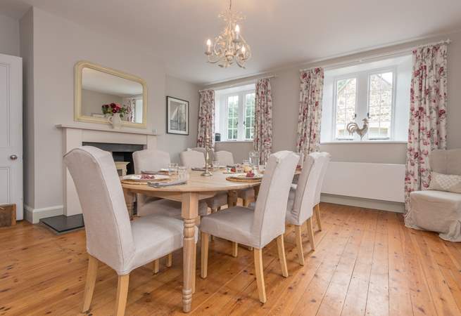 Dining room with wooden floor, table set for six, fireplace, two windows with floral curtains.