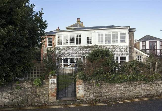 Stone house with white sunroom, behind a stone wall with a gate. Overgrown greenery in front.
