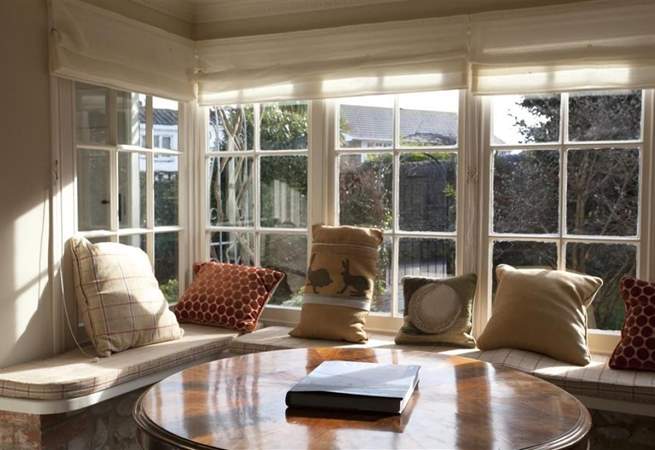 Bay window with built-in seating, cushions, and round table. Sunlight streams in, illuminating the room and exterior trees.