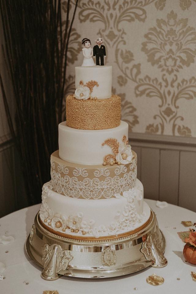 Five-tiered wedding cake with gold and white accents, topped with a bride and groom figurine, on a silver stand.