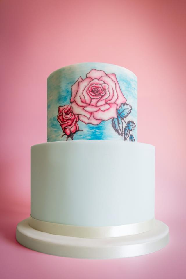 Two-tier cake with light blue frosting. Top tier has a watercolor-effect blue background and hand-drawn roses.