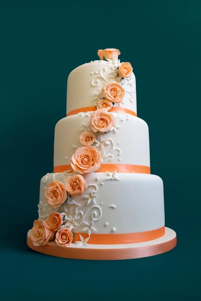 Three-tiered white cake with orange roses and ribbon, on a green background.