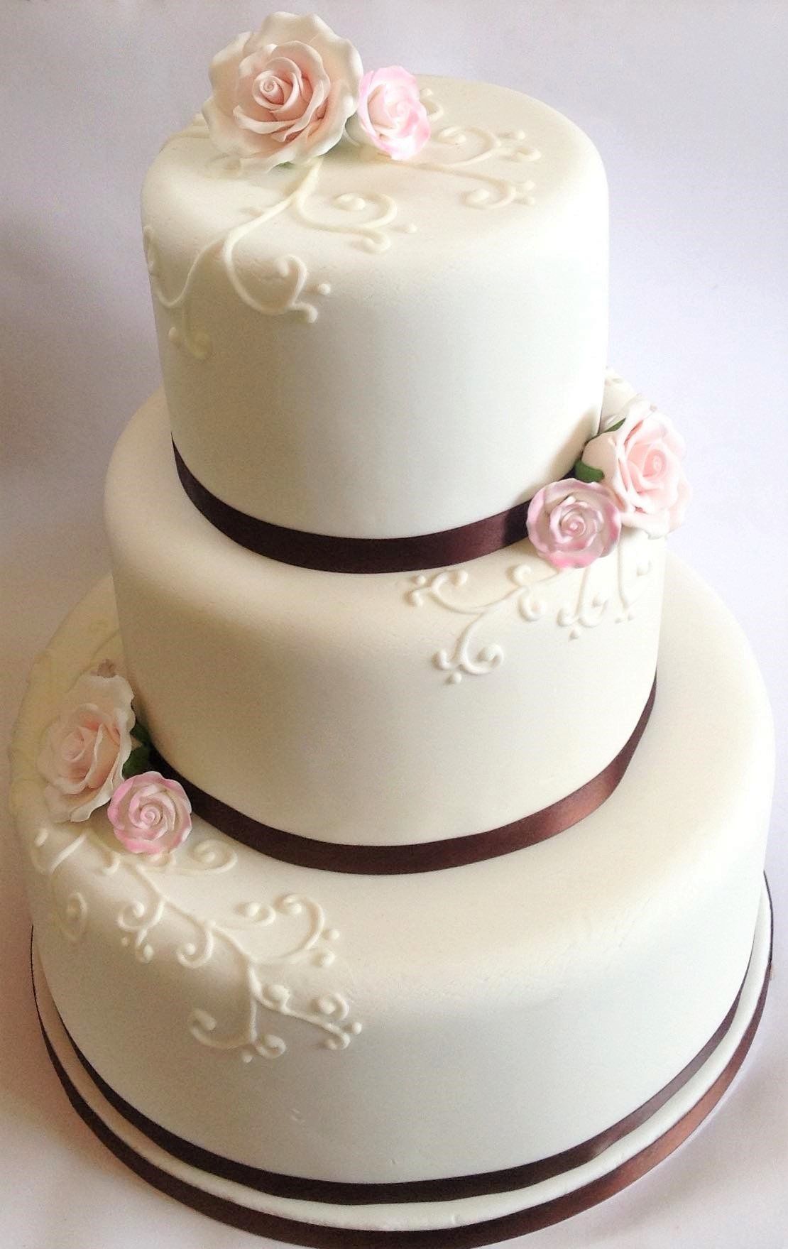 Three-tiered white cake with brown ribbon, pink roses, and white scrollwork.