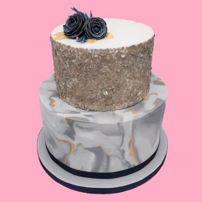 Two-tiered cake with marble and glitter icing, dark blue flowers on top, and a pink background.