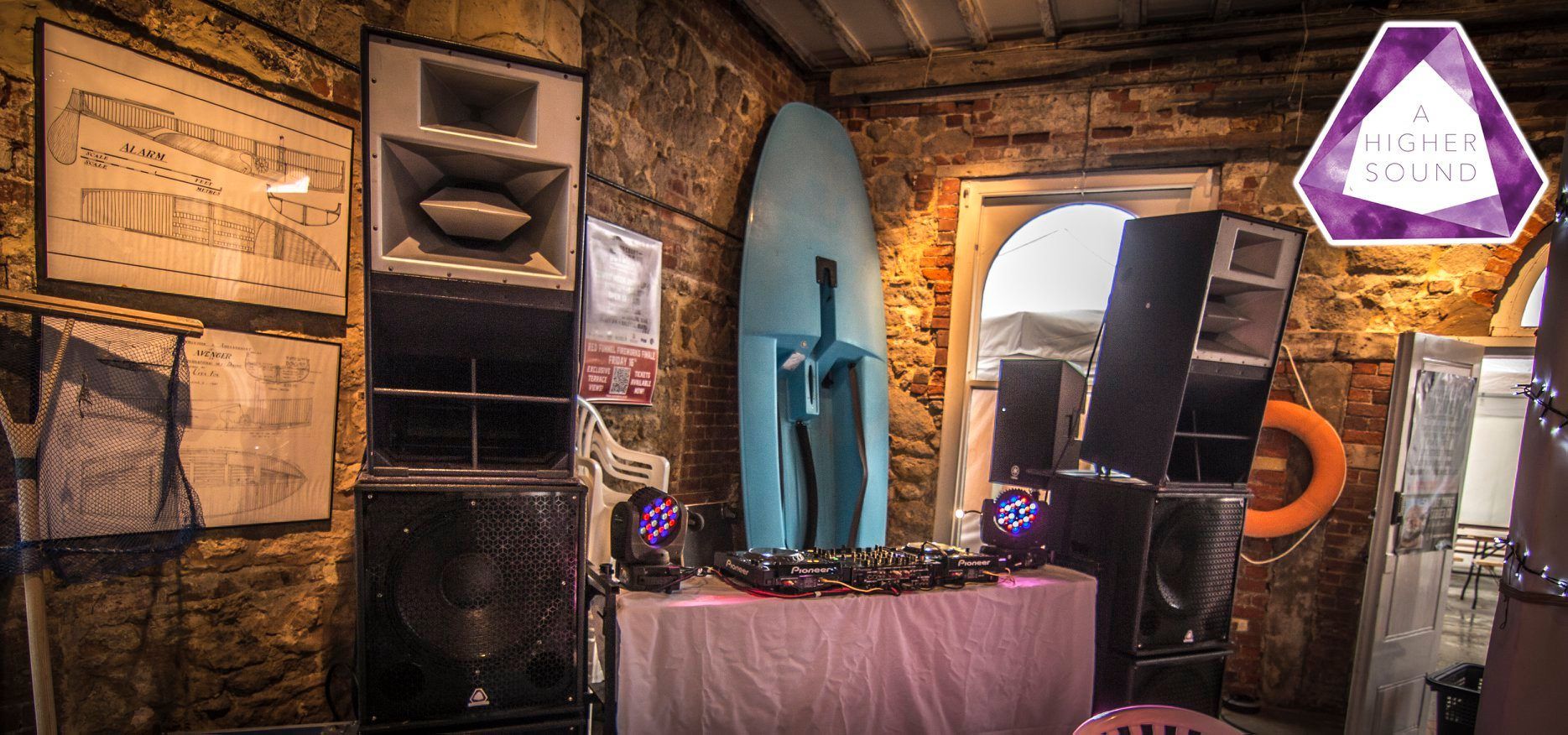 A room with DJ equipment: speakers, a surfboard, and a table. Brick walls and an arched doorway are also present.