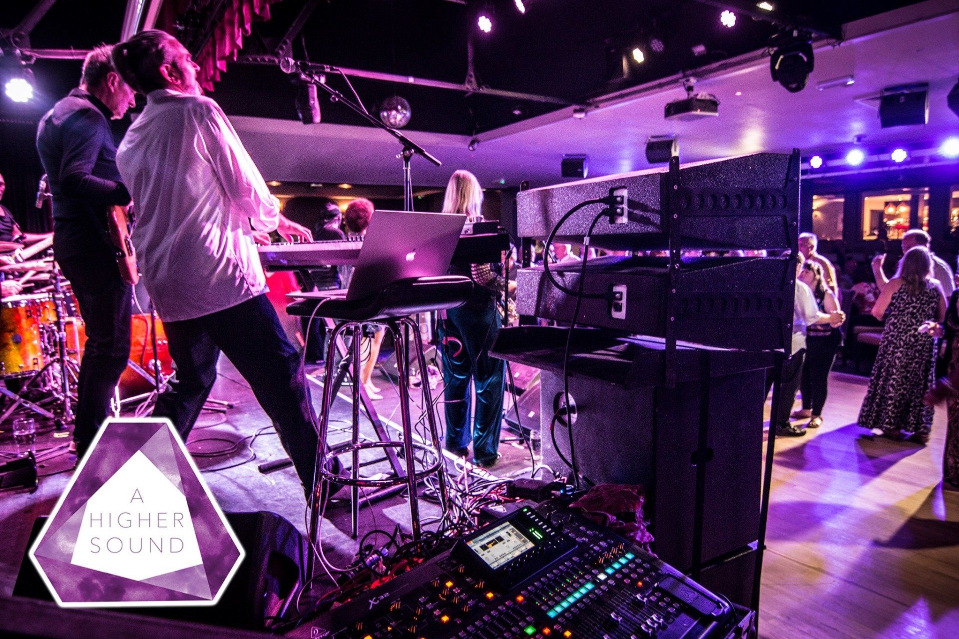 Band performing live on stage with soundboard in foreground. People dance nearby. Stage lit with purple and white lights.