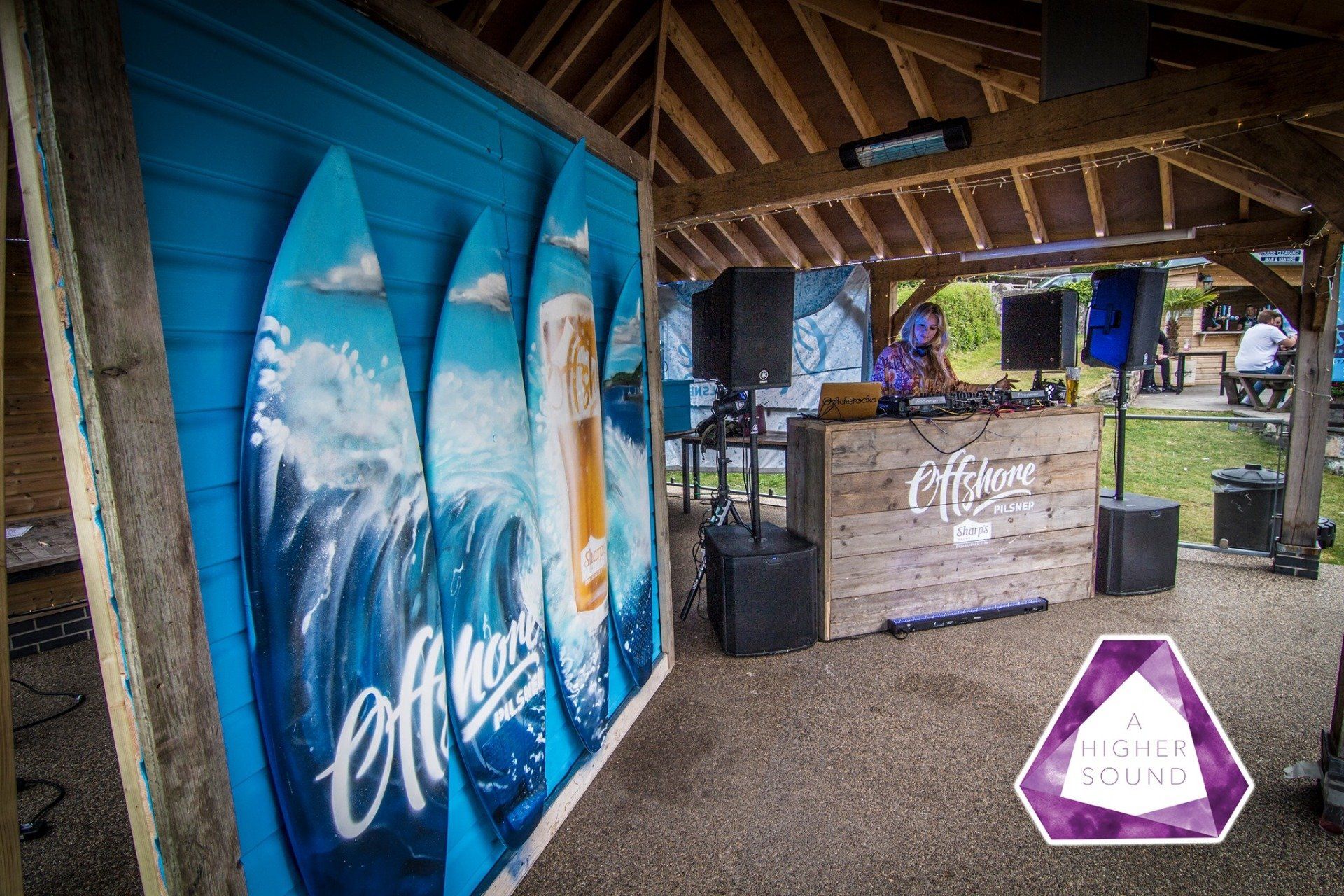 Outdoor bar with surfboards art, DJ, and stage under a wooden structure.