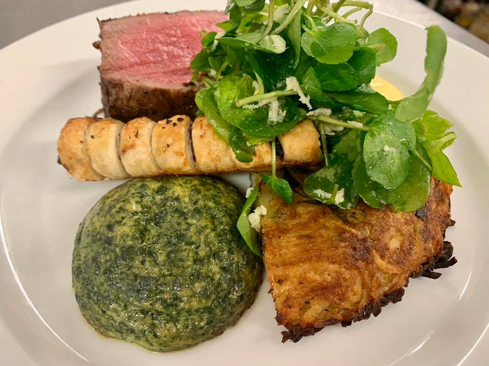 Plate with steak, puff pastry roll, spinach, potato cake, and watercress salad.