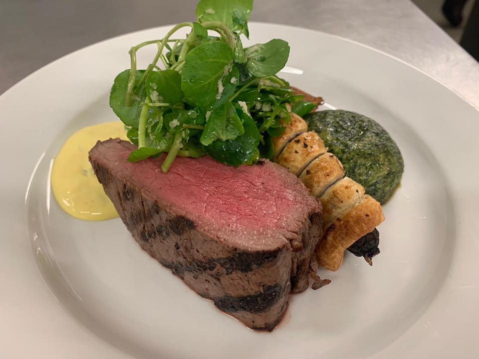 Sliced steak with greens, pastry, and sauce on a white plate.