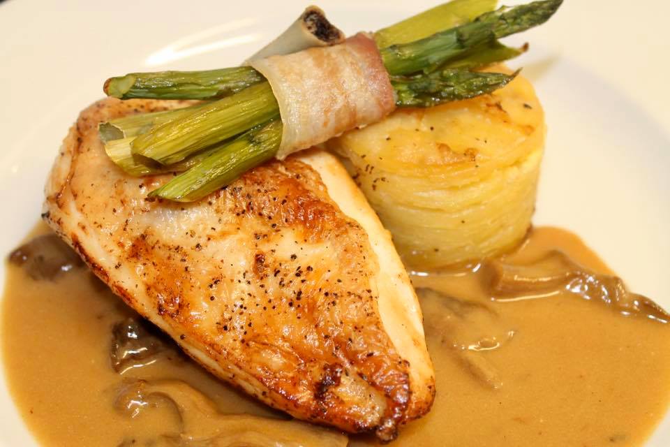 Chicken breast with mushroom sauce, asparagus wrapped in bacon, and potato tower on a white plate.
