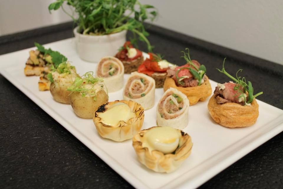 Tray of appetizers: mini quiches, wraps, potatoes, and bread topped with various fillings, with a small plant.
