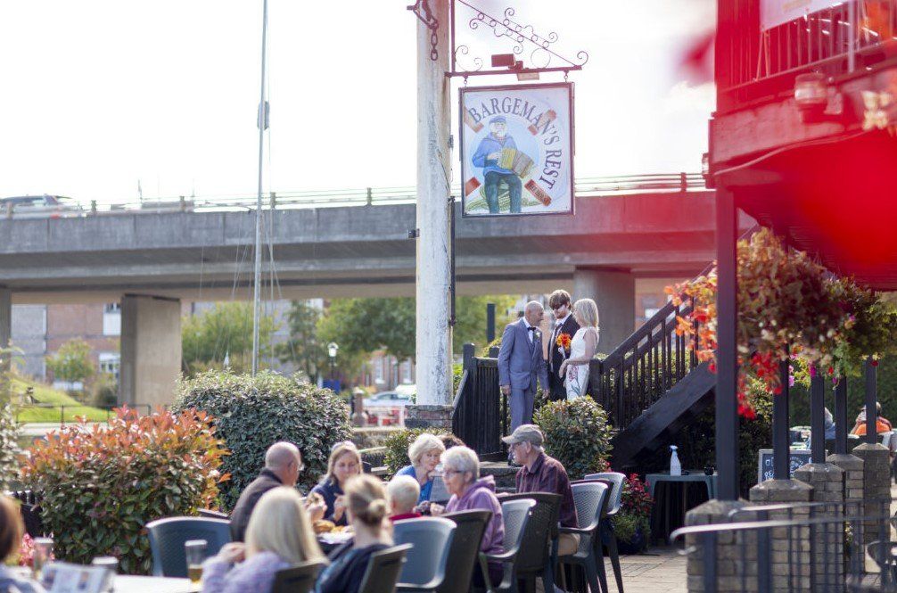 People seated outdoors at tables under the sign for a pub. A bridge is in the background.