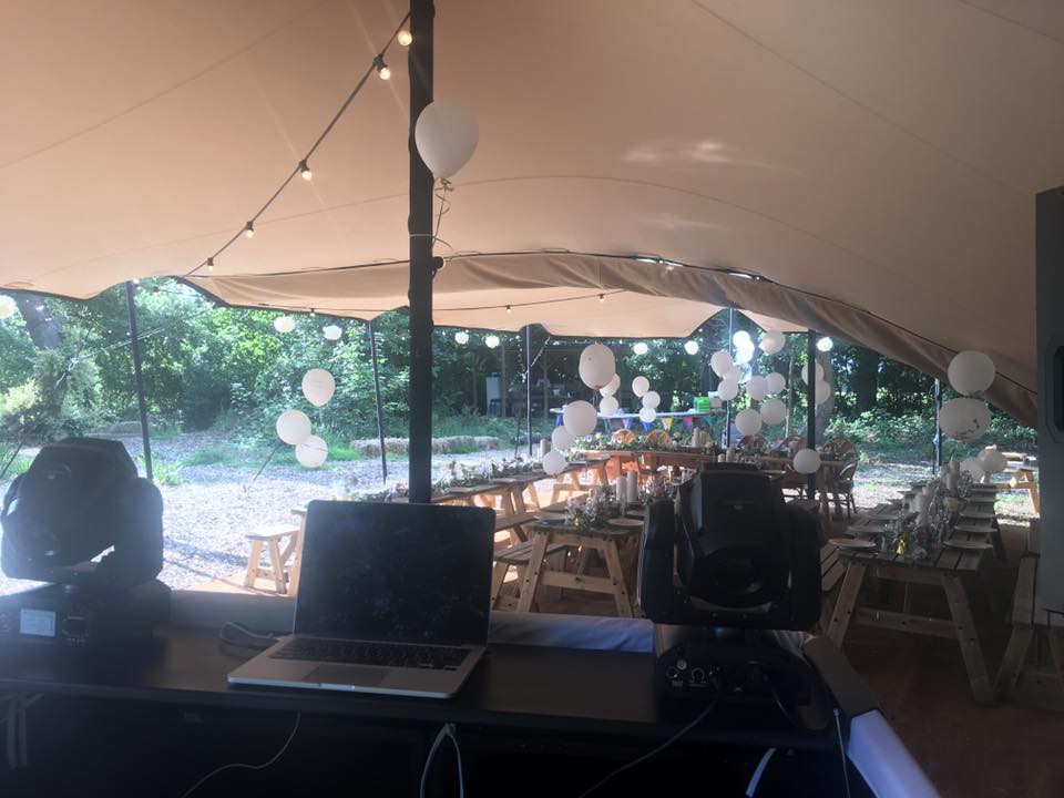 DJ setup under a tent at a party, with tables, balloons, and trees in the background.