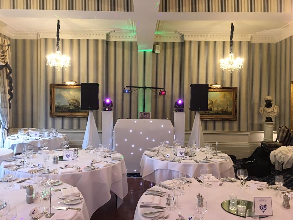 A banquet hall set for an event with round tables, a DJ setup, and decorative lighting.