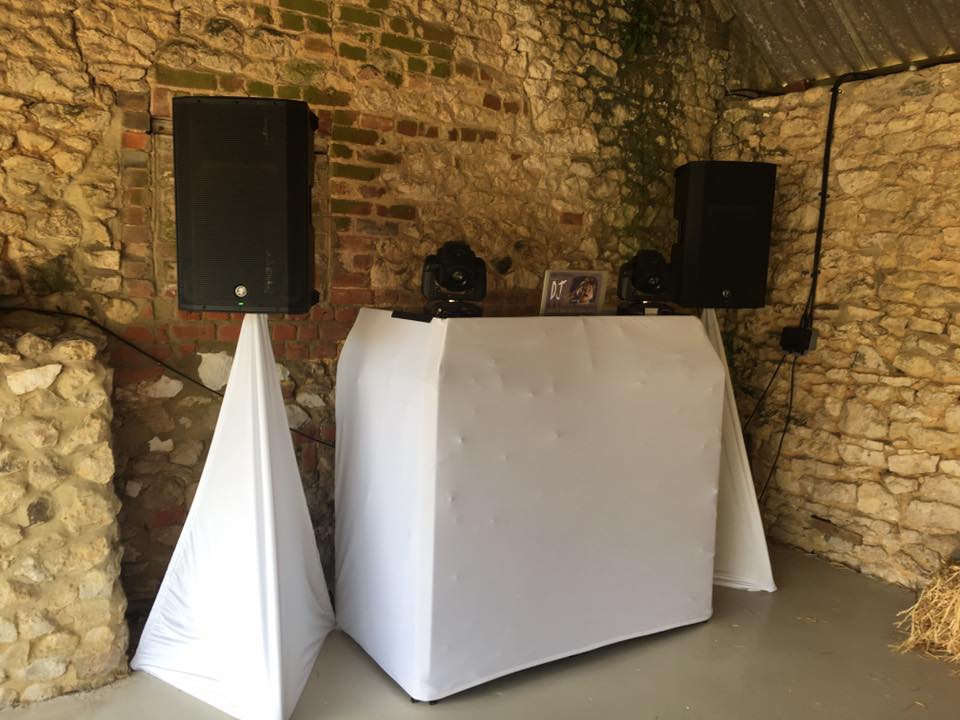DJ setup in a rustic venue: Speakers on stands, covered DJ table, and lighting against a brick and stone wall.