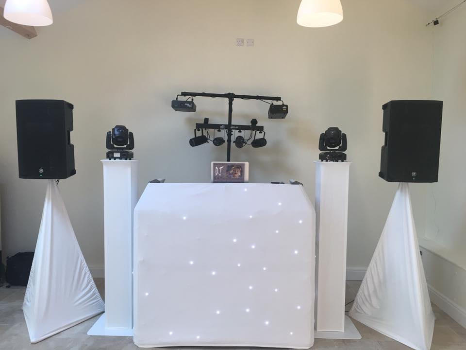 DJ setup: White facade with lights, speakers on stands, lighting truss, and two moving head lights.