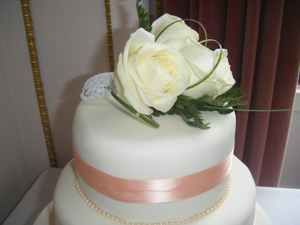Wedding cake with white frosting, pink ribbon, and white rose floral arrangement on top.