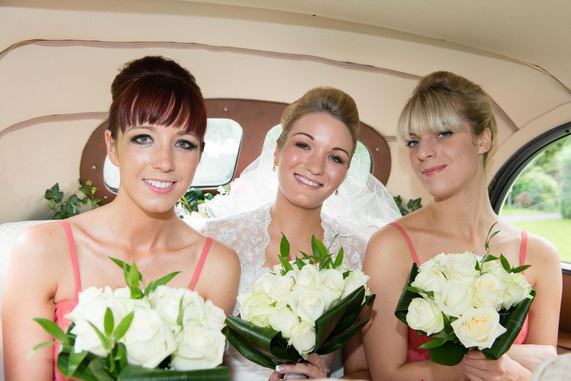 Bride and bridesmaids in a car, holding white bouquets, smiling.