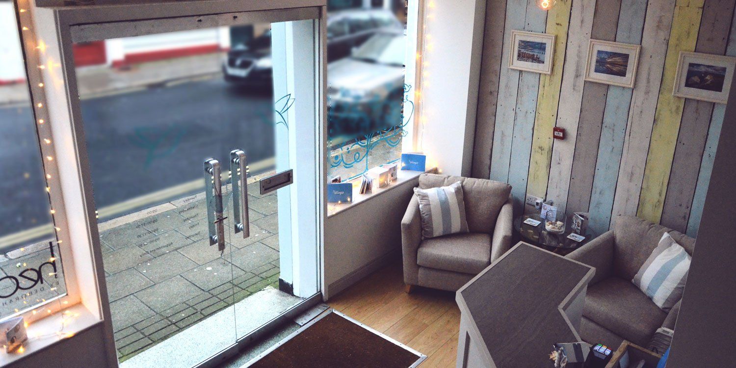 View into a shop. Glass door, waiting area with chairs, decorative wall, street visible outside.