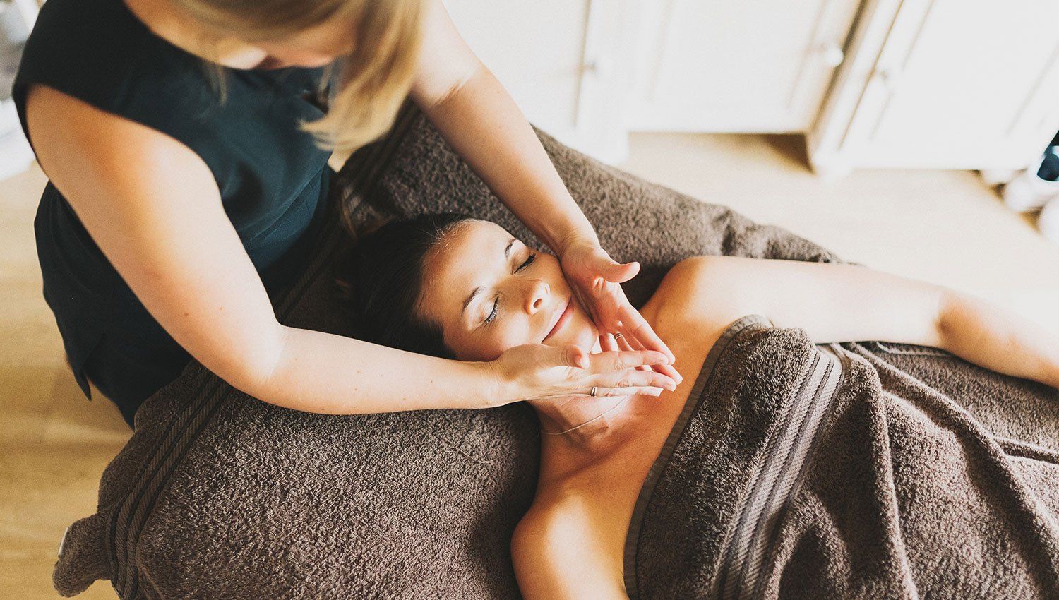 Woman receiving a facial massage, face cradled by another person's hands. Brown spa bed and towel.