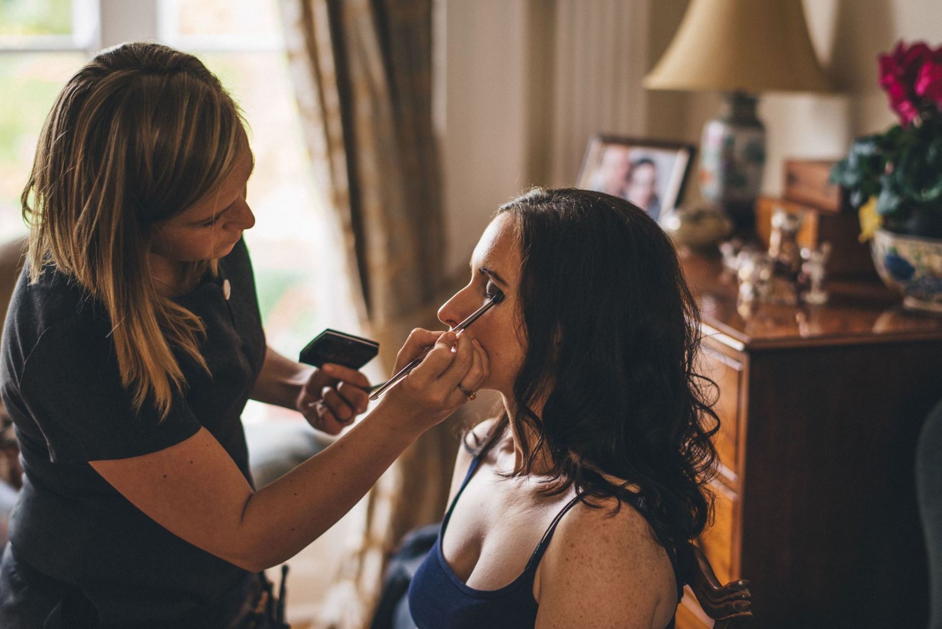 Makeup artist applying eye shadow to a woman in a room, using a brush.