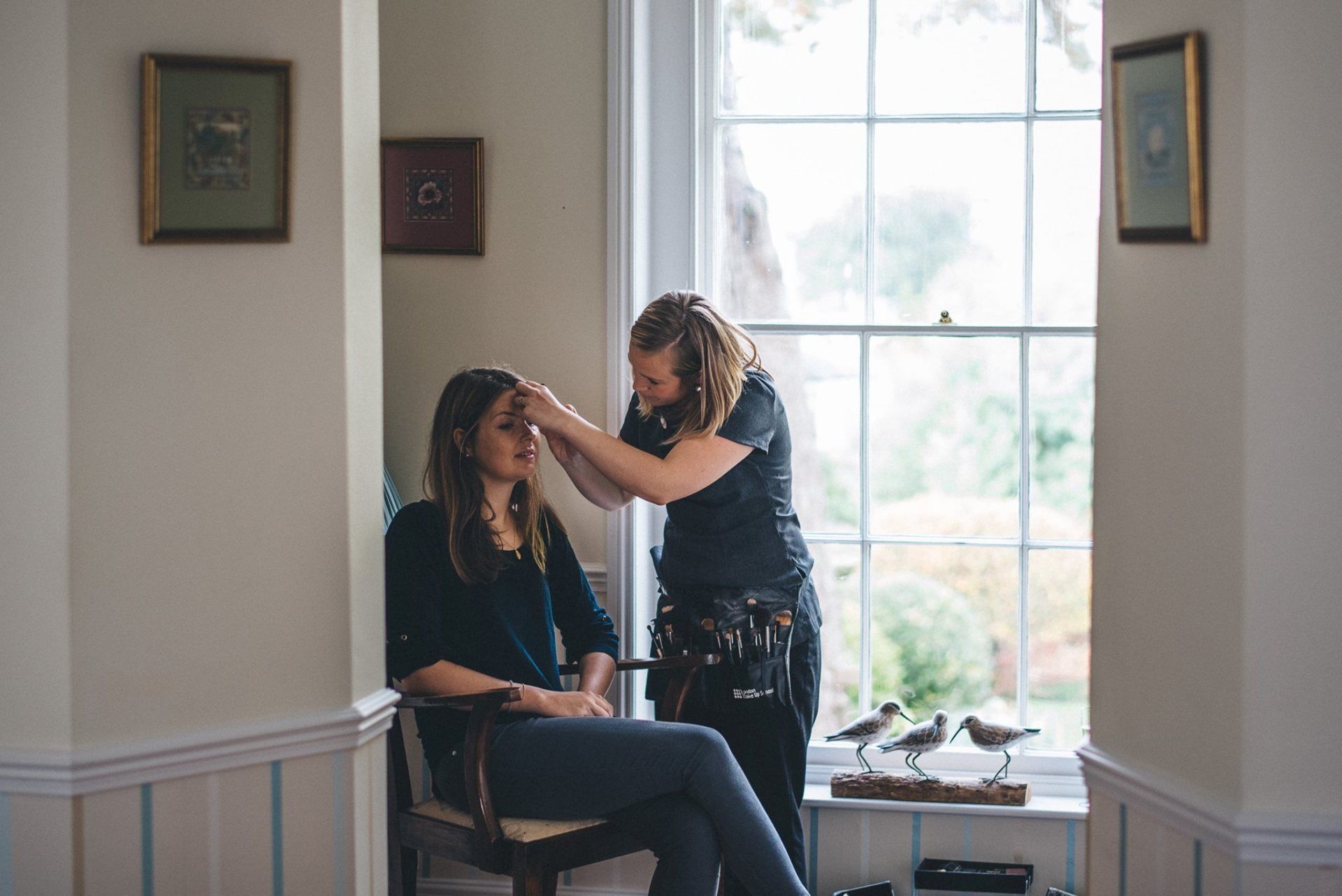 Makeup artist applying makeup to a seated person by a window in a room.