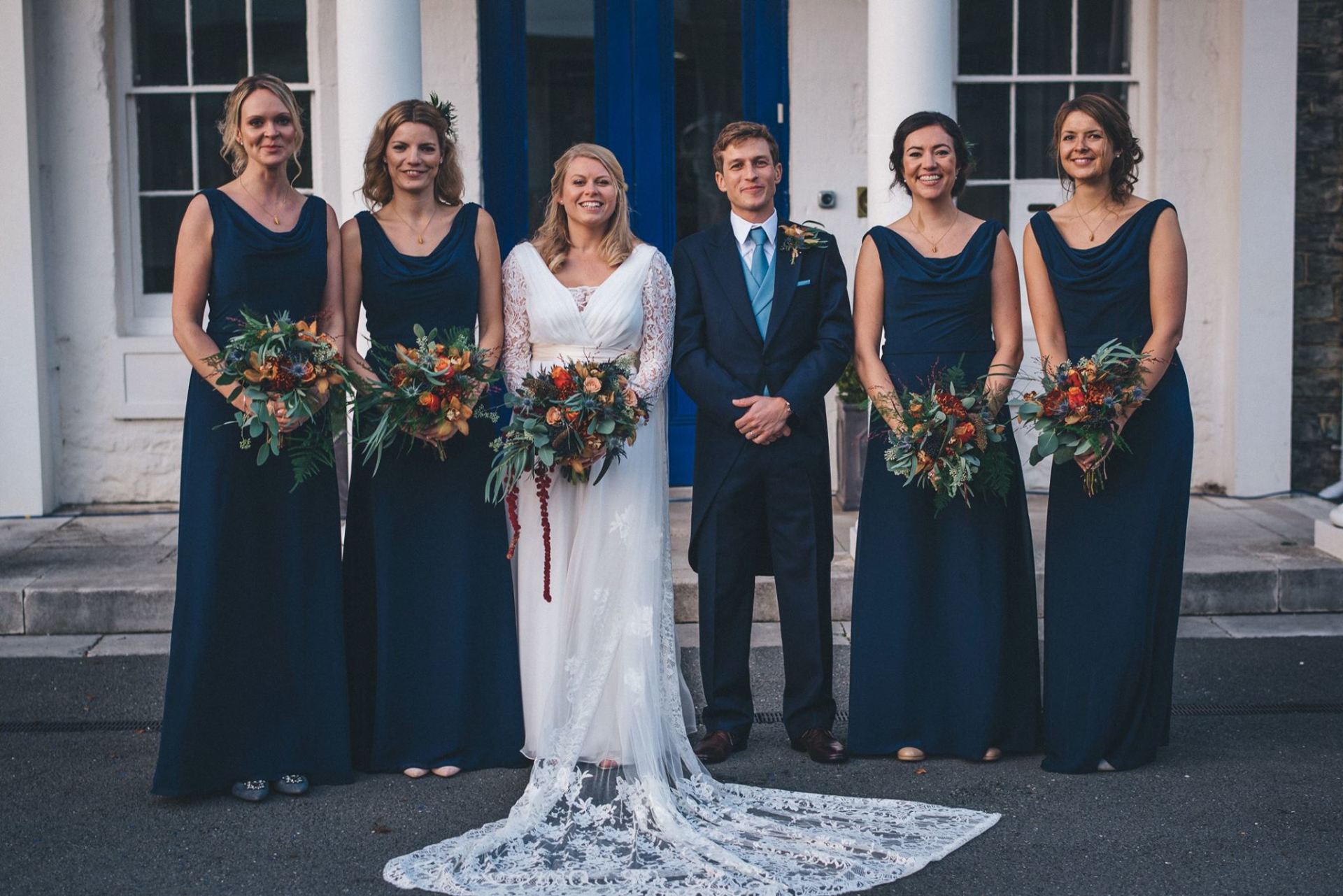 Bride and groom with bridesmaids, posing outside a building. Bride in white dress, groom in suit, bridesmaids in navy dresses holding bouquets.