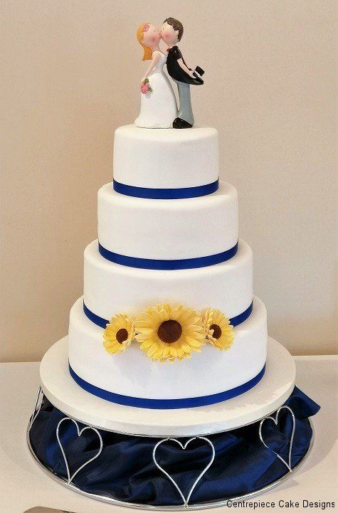 Four-tiered white wedding cake with blue ribbon and sunflowers; cake toppers of a kissing couple.