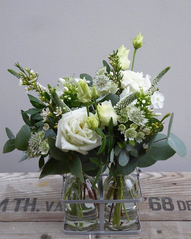 White and green floral arrangement in two glass vases held in a metal carrier, set on a wooden surface.