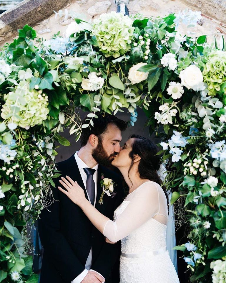 Couple kissing under a floral archway. Man in suit, woman in white dress, both in an outdoor setting.