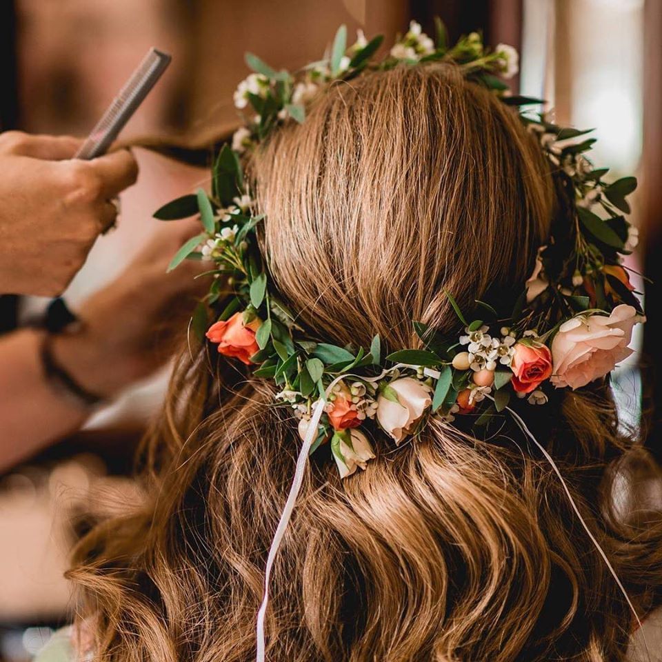 Person with a flower crown, receiving hairstyling. Hair is wavy and brown.