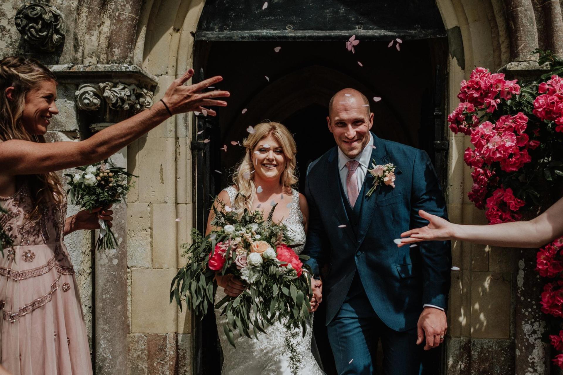 Newly married couple exits a stone church, showered with confetti. Guests throw petals; vibrant flowers frame the doorway.