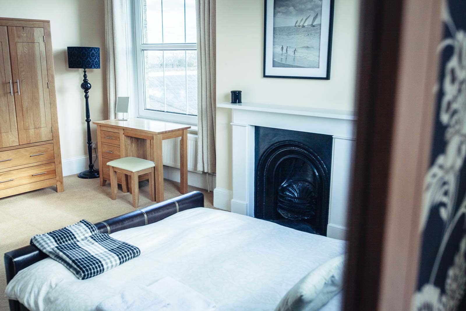 Bedroom with bed, desk, fireplace, and wooden wardrobe. Light from the window.