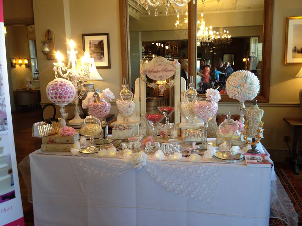 Candy buffet with various pink and white treats, displayed on a table with a sign, in a room with a large mirror.