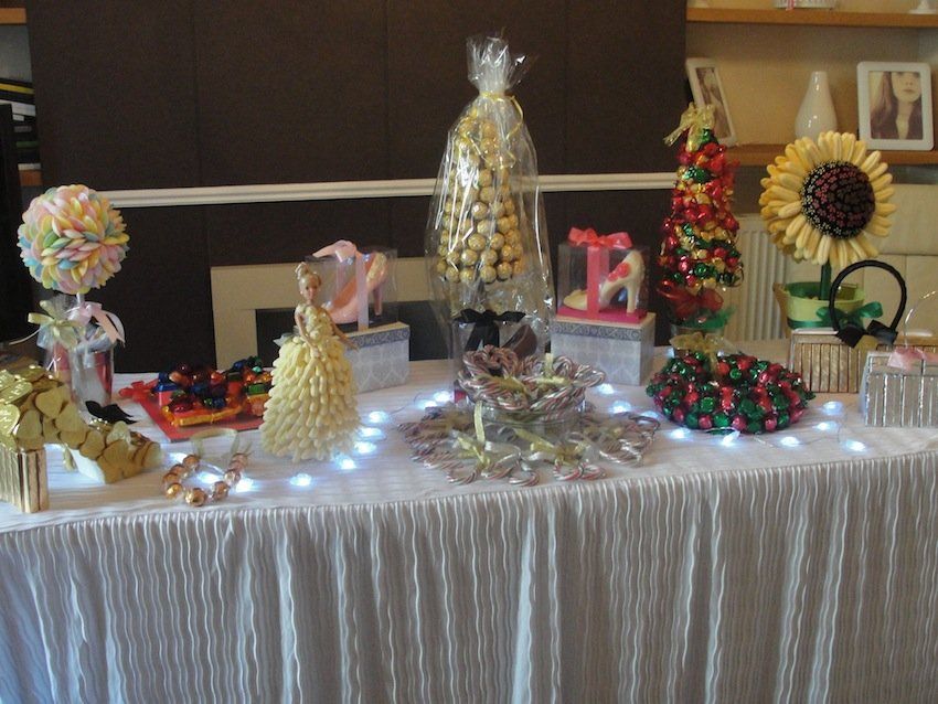 A dessert table with various sweet treats, including a sunflower, trees, and candies. White tablecloth with string lights.