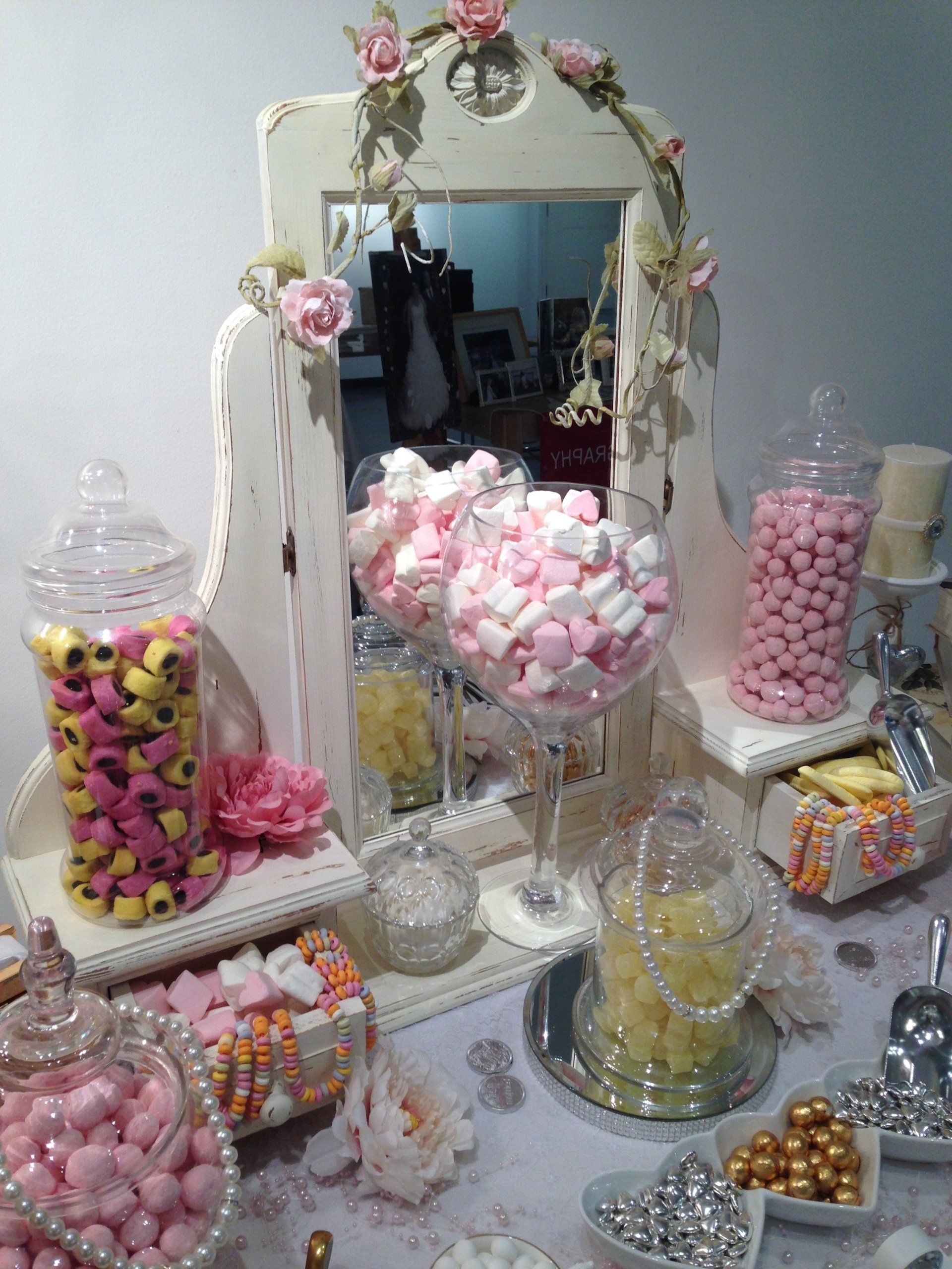 Candy buffet with jars of pink and yellow treats arranged around a mirror decorated with flowers.
