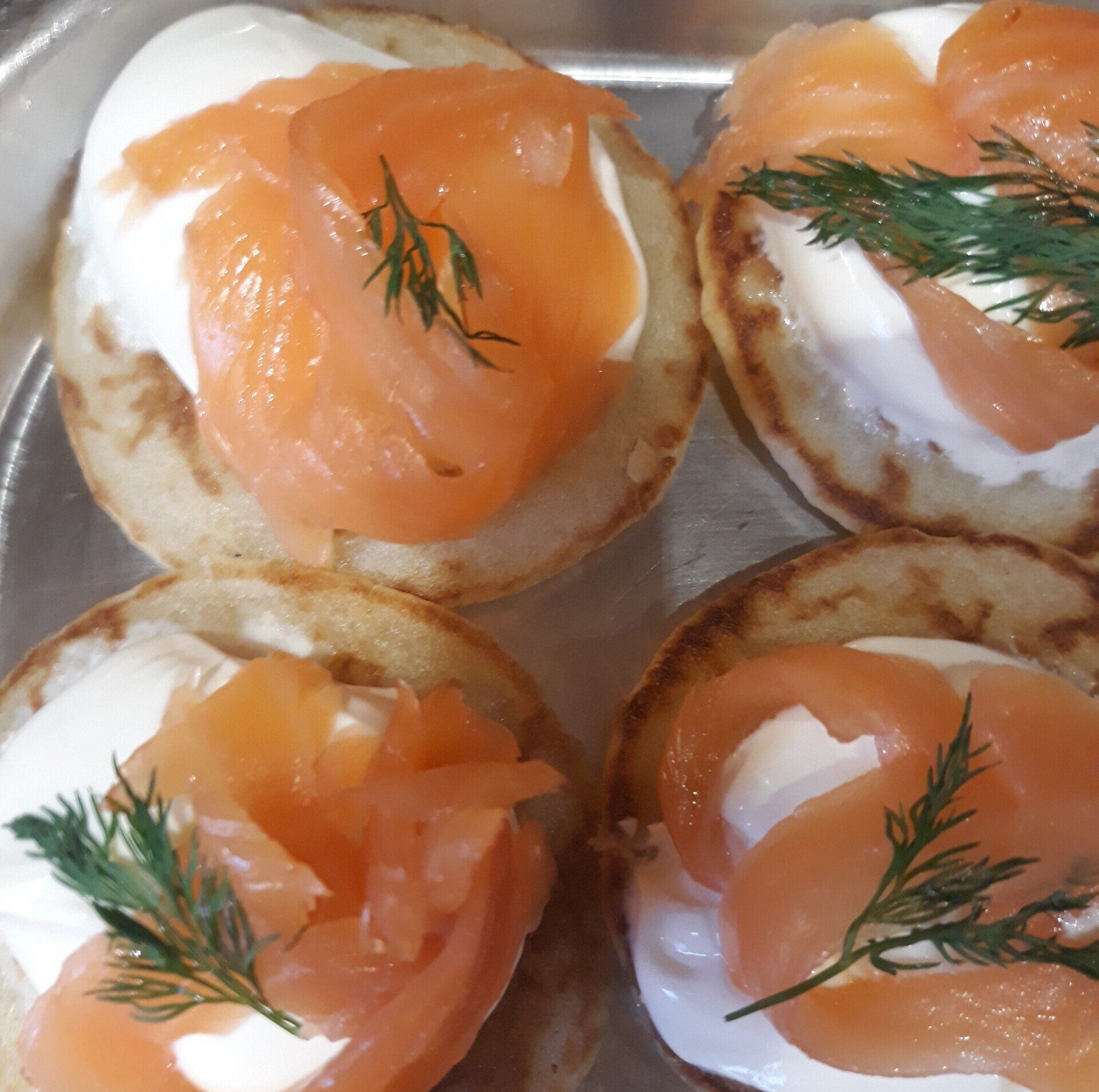 Four blinis topped with cream cheese, smoked salmon, and dill sprigs on a metal tray.