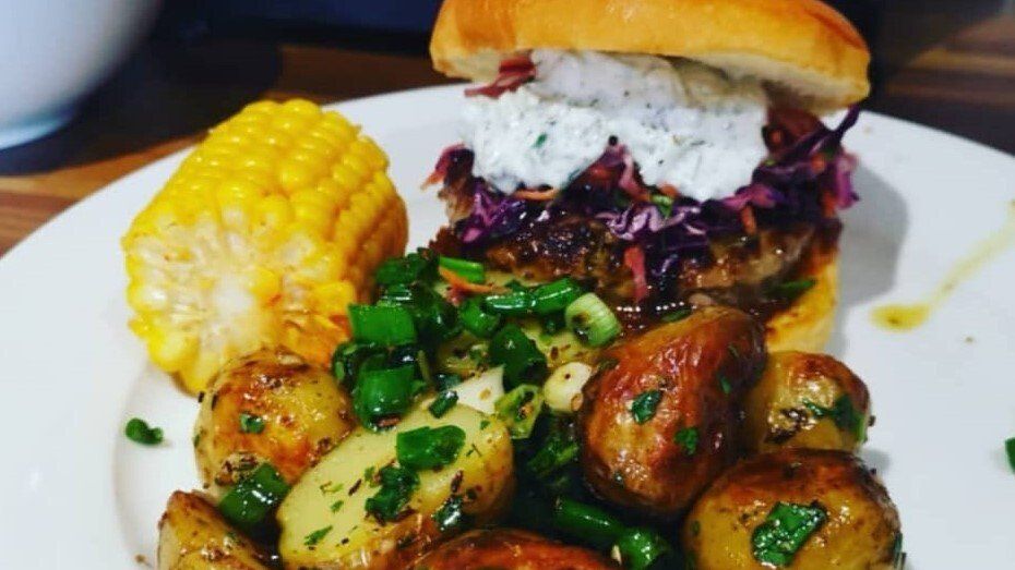 Burger on bun, topped with slaw and sauce, beside corn on the cob and potatoes with green herbs on white plate.