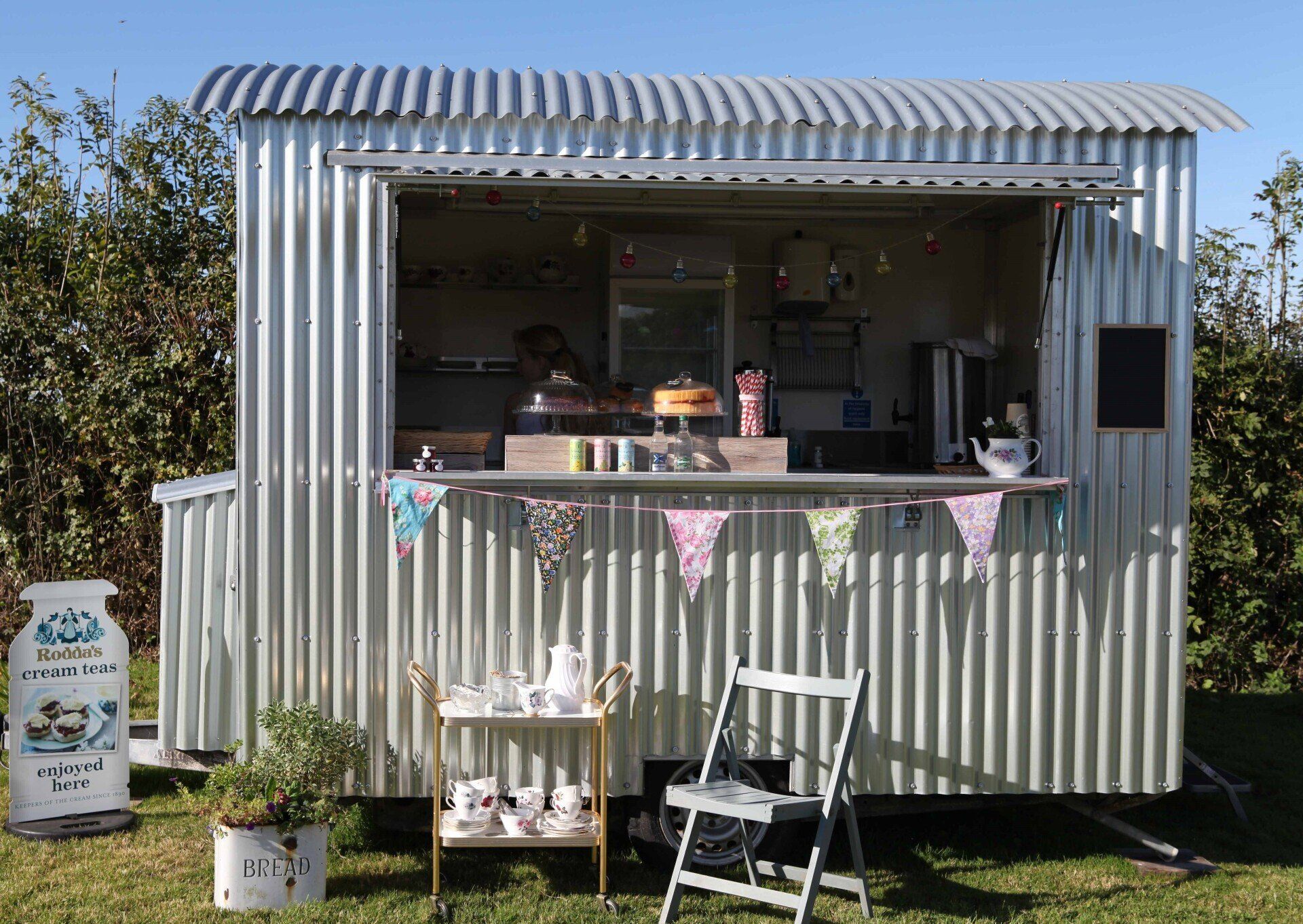 Corrugated metal food stand with open window, decorated with bunting, table and chair.