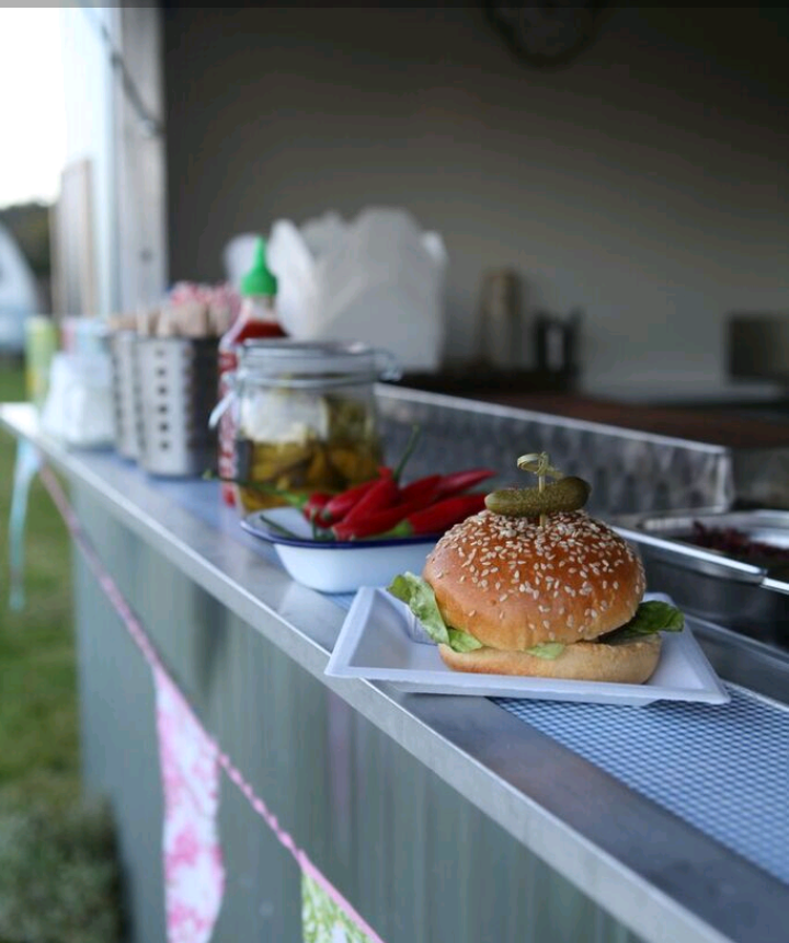 Burger on a food truck counter, with toppings, next to peppers and condiments.