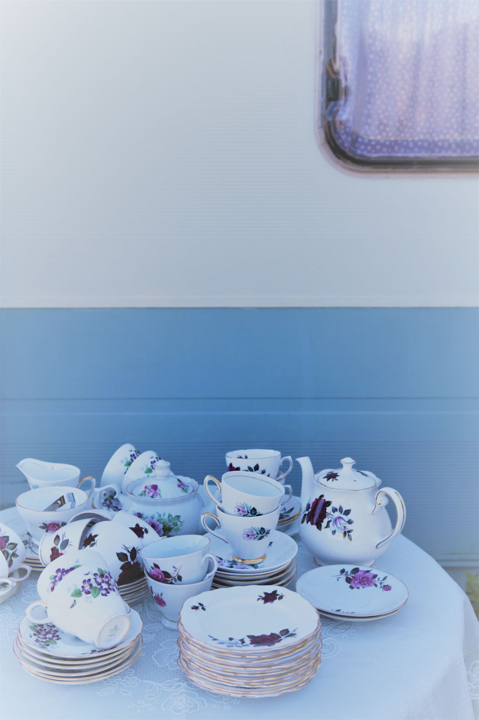 Teapot and teacups with purple floral pattern on a white tablecloth, near a blue and white wall.