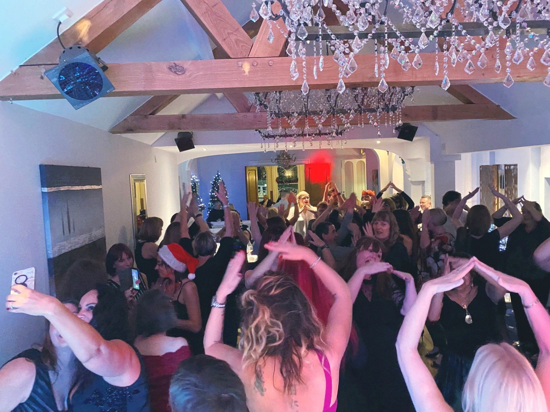 People dancing with raised arms at a festive party in a room with a chandelier.
