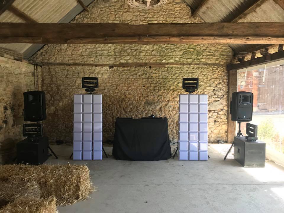 DJ setup in a rustic barn with speakers, lights, and a table covered in black fabric.