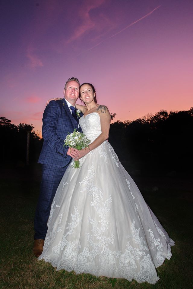 Bride and groom pose outdoors, holding bouquet, beneath a colorful sunset. Groom in blue suit, bride in white gown.