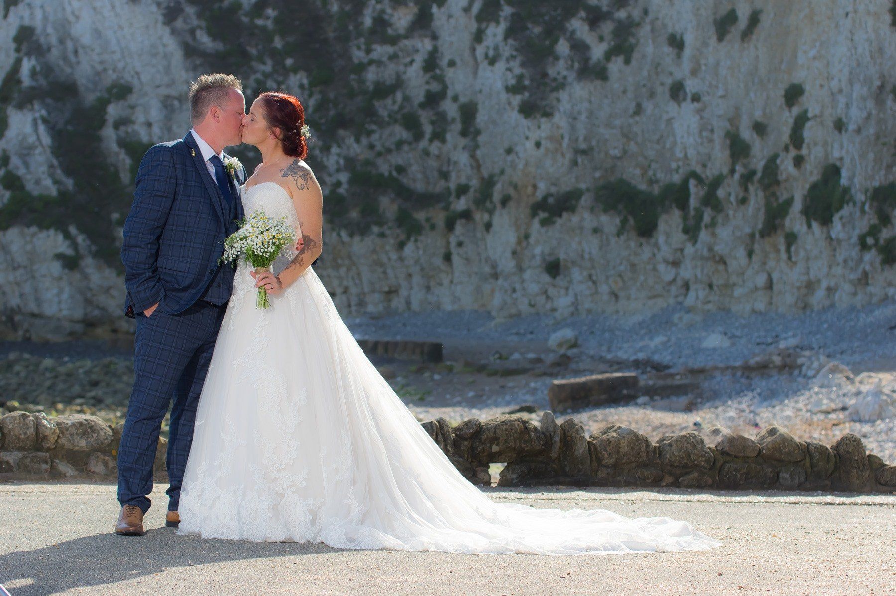 Couple kissing in wedding attire; white dress, blue suit; cliffs in background.