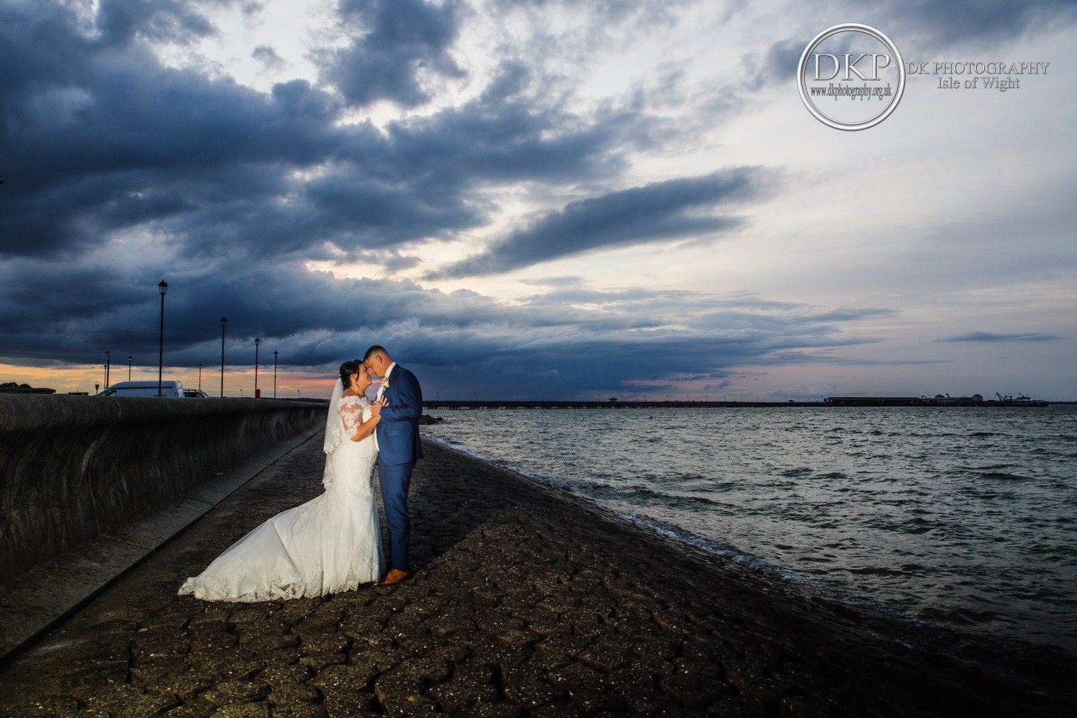 Bride and groom embrace on a stone pier near water under a dramatic, cloudy sky at dusk.